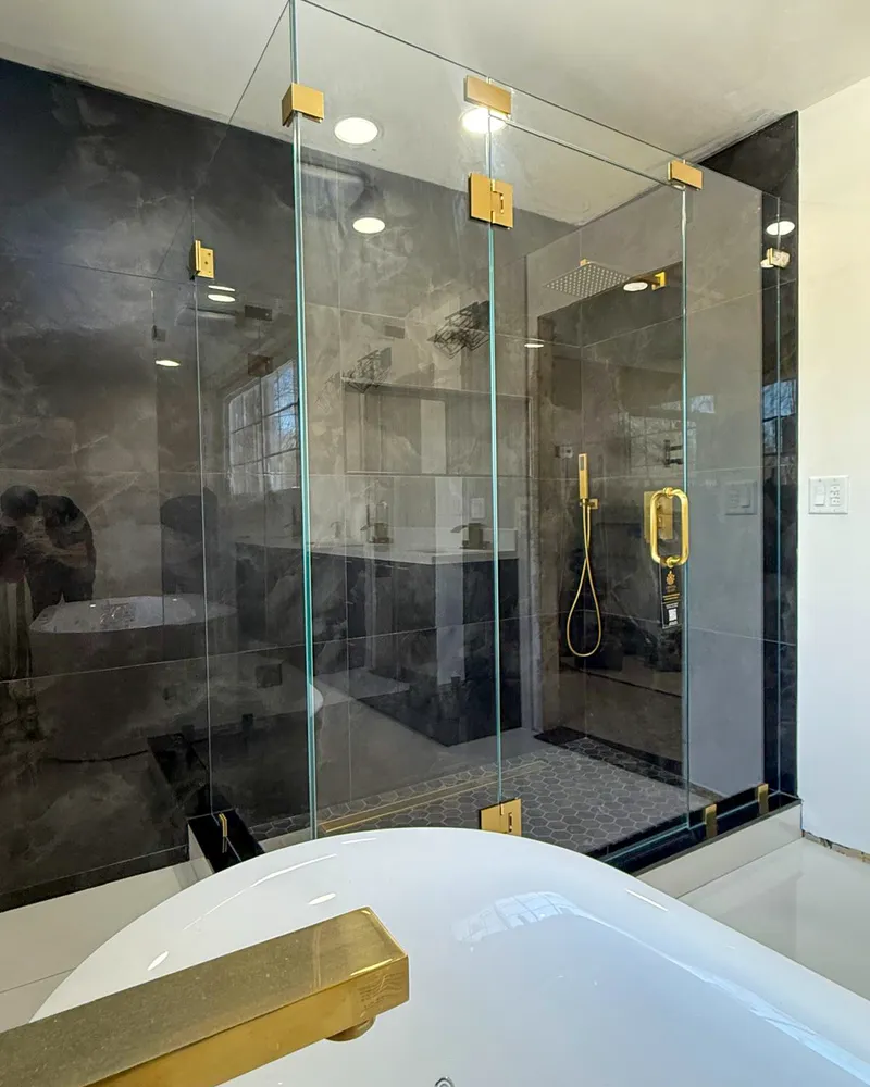 Frameless enclosure with brushed gold hardware against dark veined stone, beside a freestanding tub